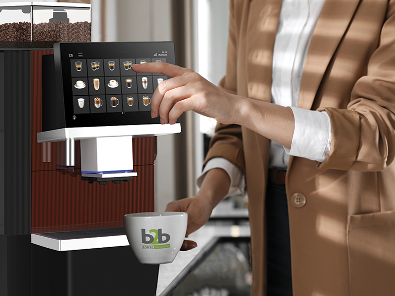 A professional in a tan blazer uses a finger to select a drink on the large digital touchscreen of a premium bean-to-cup coffee machine while holding a white ceramic cup featuring the "b2b Coffee Solutions" logo.
