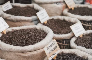 A collection of burlap sacks filled with different types of roasted coffee beans, each with a small handwritten sign identifying the origin and roast type.