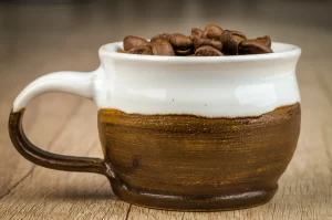 A close-up side view of a rustic, handmade ceramic coffee mug filled with roasted coffee beans, resting on a light wood-grain surface.