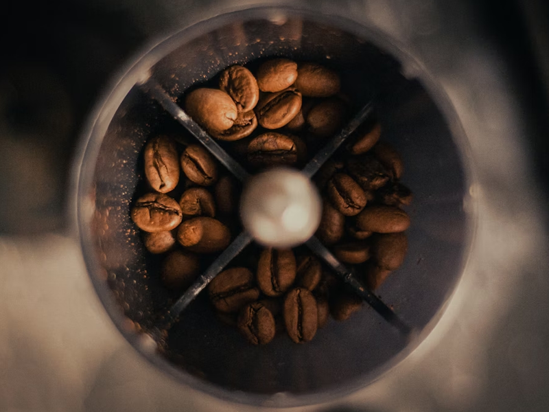 A dramatic overhead close-up shot of dark roasted Uasal coffee beans inside a circular grinder hopper, showing the mechanical divider and the oily texture of the fresh beans.
