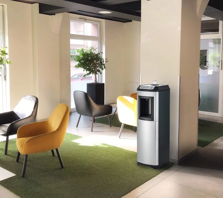 A sleek, Oasis KaliX Water Cooler situated in a modern office lobby with yellow and grey armchairs and green rugs.