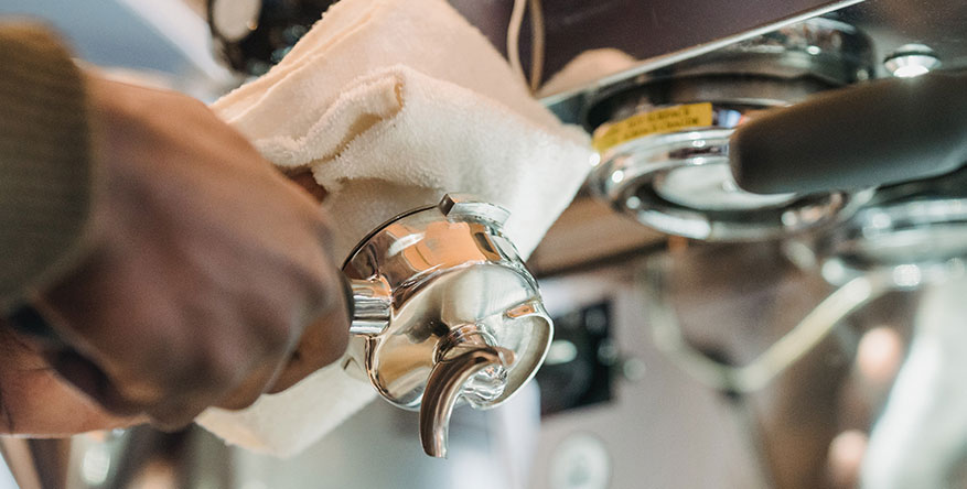A close-up view of a person's hand using a white cloth to wipe the metal underside of an espresso machine portafilter.