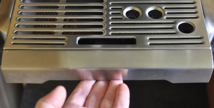 A hand reaches toward the underside of a stainless steel drip tray on an espresso machine.