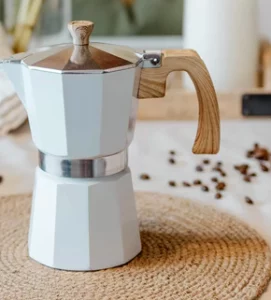 A professional product shot of a modern, white-coated Moka pot with a natural wood-grain handle and knob, resting on a woven jute coaster with scattered coffee beans in the background.