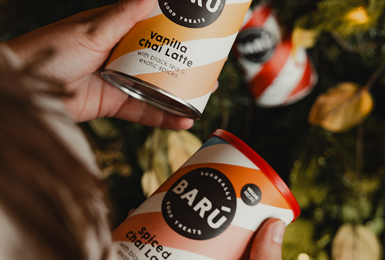Close-up of hands holding two BARÚ canisters—Vanilla Chai Latte and Spiced Chai Latte—against a background of festive greenery and warm lights.