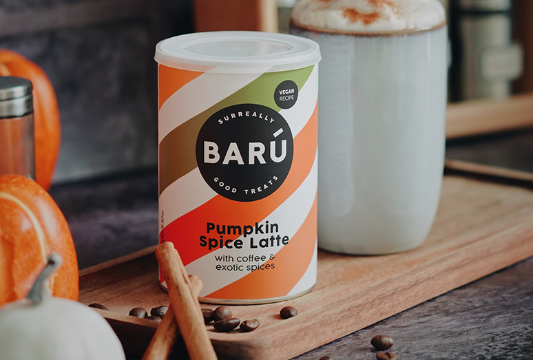 A professional product shot of a BARÚ Pumpkin Spice Latte canister on a wooden tray, accompanied by cinnamon sticks, coffee beans, and a tall latte glass in a seasonal autumnal setting.