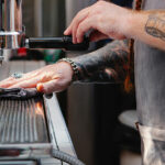 A close-up of a barista with tattooed arms wearing a grey apron, carefully cleaning the drip tray of a professional espresso machine with a grey microfiber cloth.