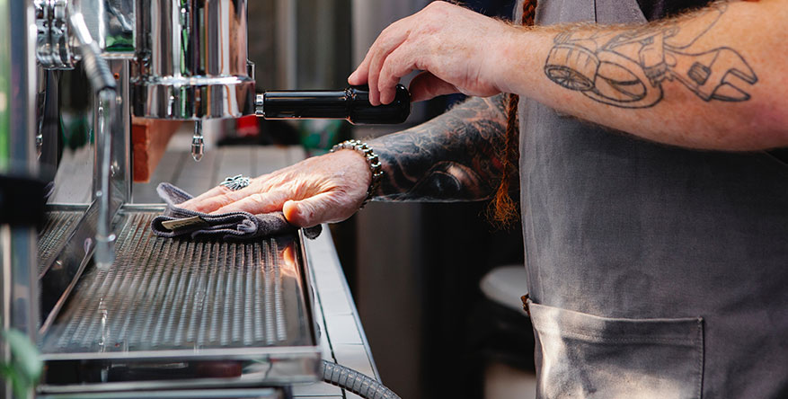 A close-up of a barista with tattooed arms wearing a grey apron, carefully cleaning the drip tray of a professional espresso machine with a grey microfiber cloth.