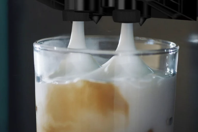 A close-up view of two spouts from a coffee machine dispensing thick, creamy frothed milk into a glass containing liquid.