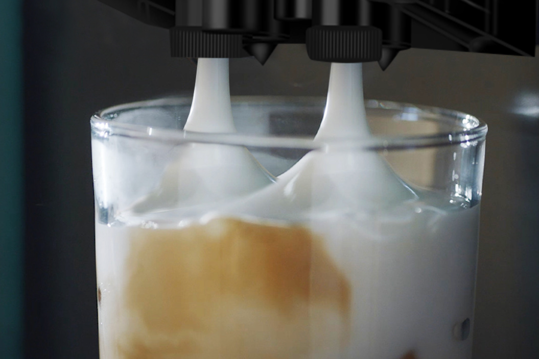 A close-up view of two spouts from a coffee machine dispensing thick, creamy frothed milk into a glass containing liquid.