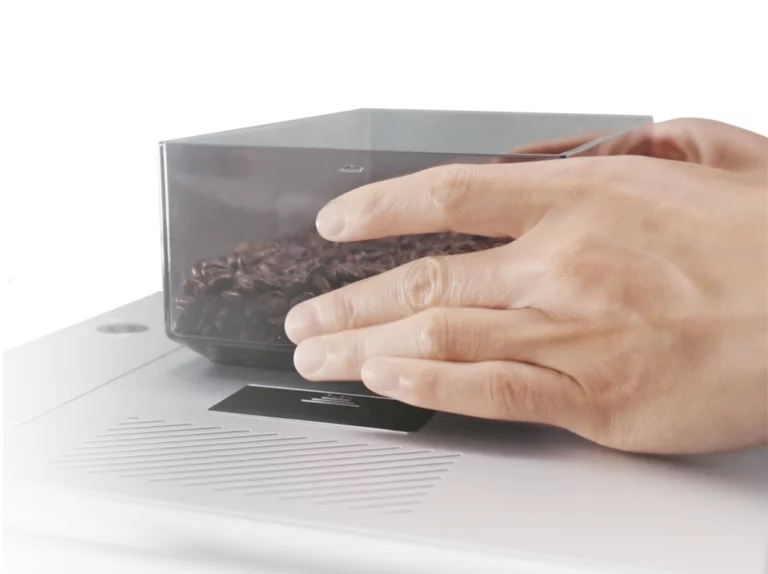 A close-up shot of hands placing a transparent container filled with dark-roasted coffee beans onto the surface of a modern, sleek, silver electronic coffee scale.