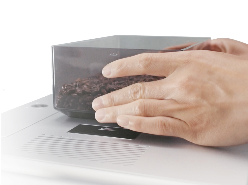 A close-up shot of hands placing a transparent container filled with dark-roasted coffee beans onto the surface of a modern, sleek, silver electronic coffee scale.