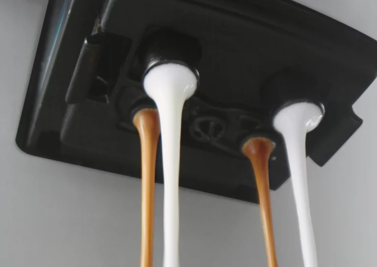 A close-up, low-angle view of a coffee machine spout dispensing two streams of dark espresso and two streams of white milk simultaneously.
