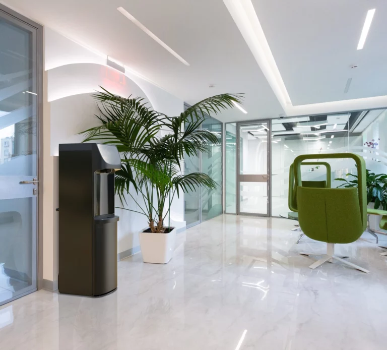 A slim, floor-standing Oasis Danu Water Cooler situated in a bright, modern office lobby with white marble floors, glass walls, and green lounge chairs.