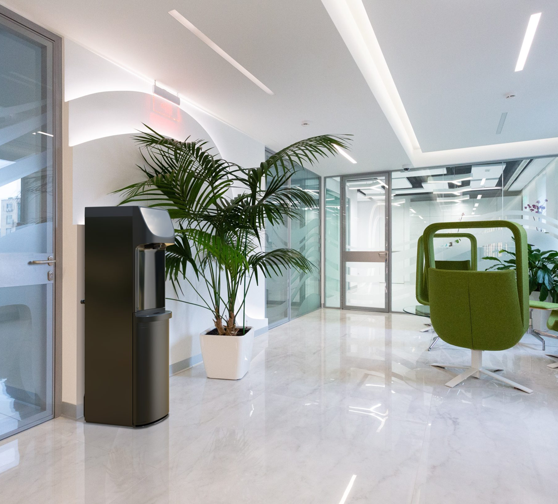 A slim, floor-standing Oasis Danu Water Cooler situated in a bright, modern office lobby with white marble floors, glass walls, and green lounge chairs.
