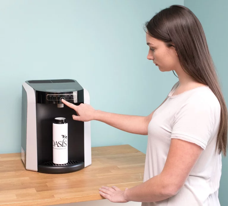A person pressing a button on the top touch panel of a Polaris Water Cooler Oasis water dispenser to fill a white reusable water bottle on a wooden table.