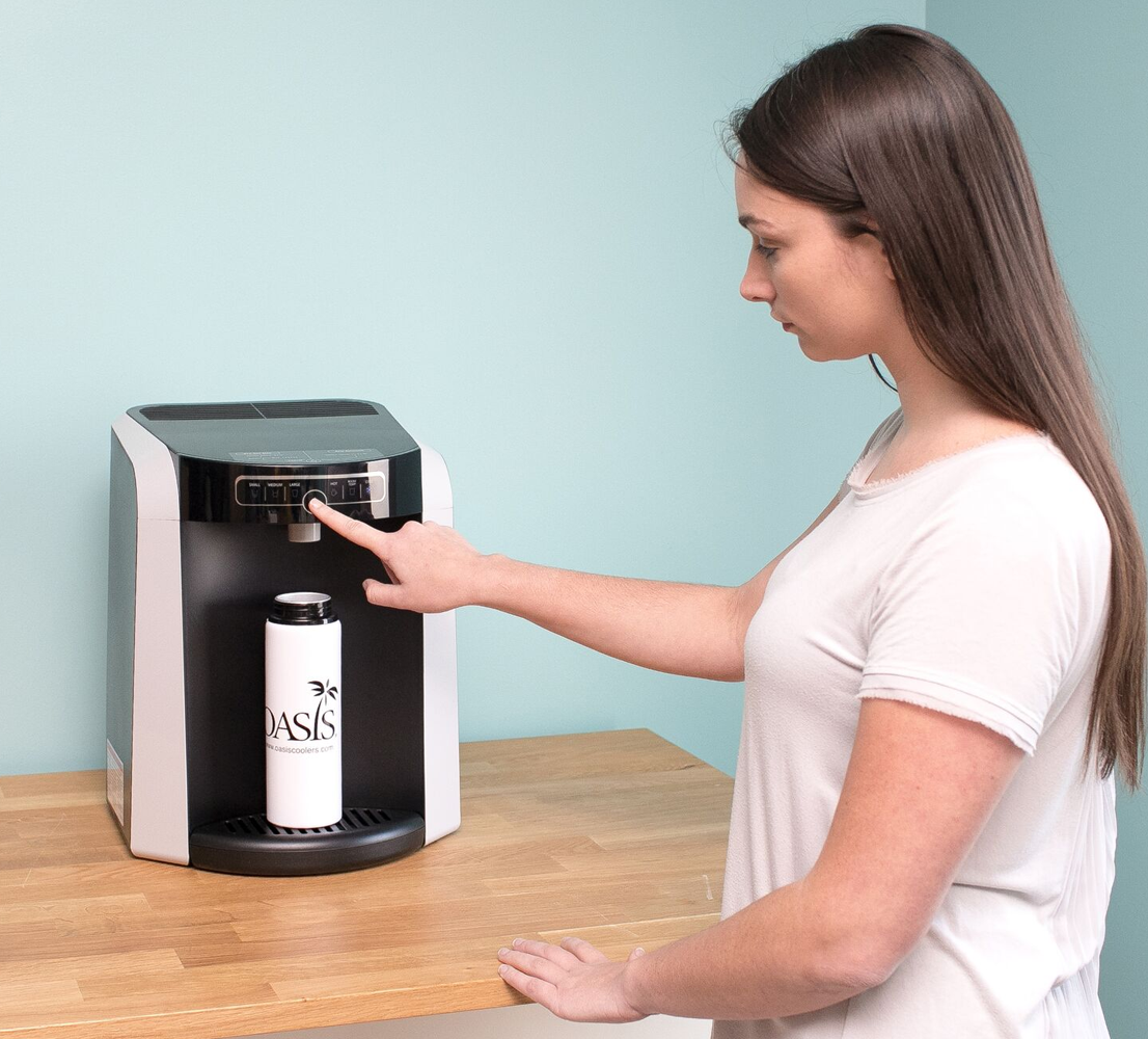 A person pressing a button on the top touch panel of a Polaris Water Cooler Oasis water dispenser to fill a white reusable water bottle on a wooden table.