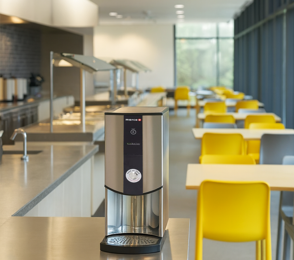 A Marco EcoBoiler water boiler on a stainless steel counter in a modern, brightly lit office canteen with yellow and grey chairs in the background.