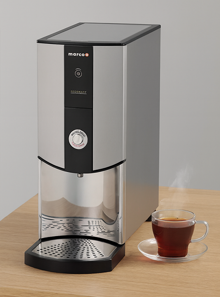 A professional studio shot of a Marco Ecoboost stainless steel water boiler on a wooden surface, next to a clear glass cup of steaming tea on a saucer.