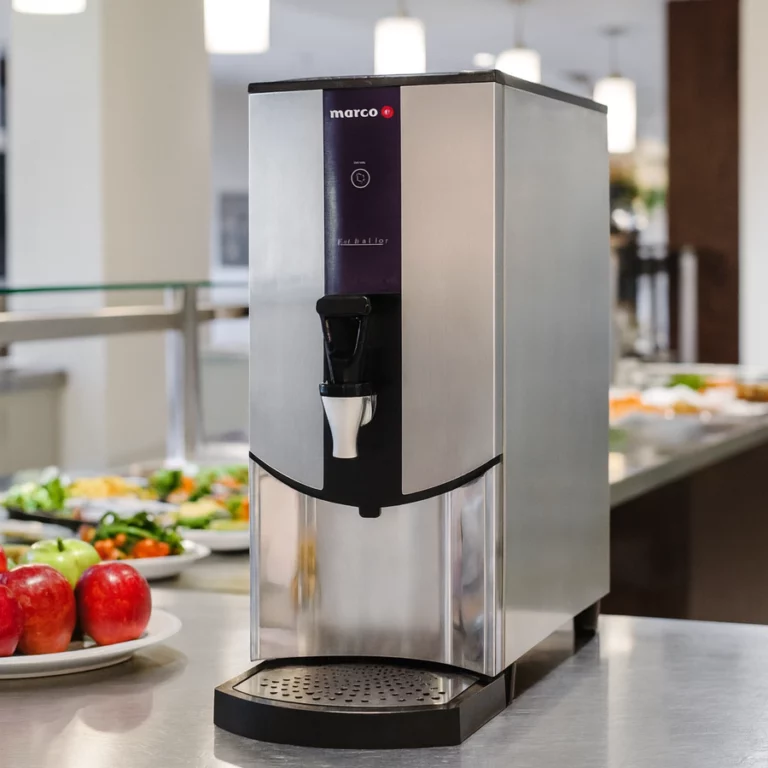 A Marco Ecoboiler on a white countertop in a bright office cafeteria, with fresh apples and salads in the foreground and blurred dining tables by a window in the background.