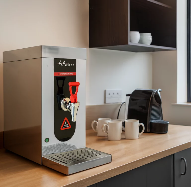 An AA First PowerSmart water boiler situated on a wooden office countertop next to white ceramic mugs and a compact espresso machine in a modern kitchen setting.