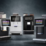 A wide-angle product lineup featuring four distinct professional bean-to-cup coffee machines—including high-capacity, white-panel, and flagship black models—all arranged against a dark, moody background with atmospheric smoke.