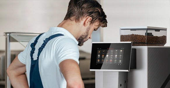 A professional technician in a white polo shirt and blue work overalls leaning in to inspect the digital touchscreen interface of a premium bean-to-cup coffee machine.